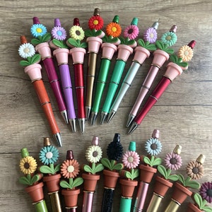 May include: A collection of 18 flower-shaped pens with pink, green, red, purple, gold, and silver barrels. The pens have a flower design with a pot and leaves. The flowers are in various colors, including pink, yellow, blue, and orange.