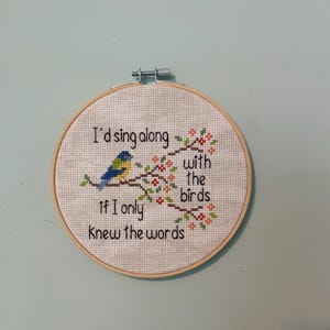 Peut inclure: Un cercle de broderie au point de croix avec le texte "I'd sing along with the birds if I only knew the words." Le motif représente un oiseau bleu et jaune perché sur une branche fleurie. Le cercle est en bois.