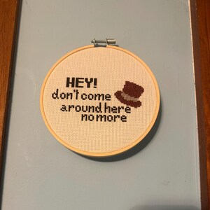 May include: A cross-stitch hoop with the text "HEY! don't come around here no more" and a small brown hat. The text and hat are stitched in dark brown thread on a cream-colored fabric, set in a wooden hoop.