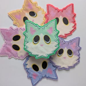 May include: A collection of six colorful cat-shaped patches. Each patch features a fluffy face with large black eyes, a small pink nose, and a white center. The patches are in shades of pink, purple, blue, green, and peach, with embroidered details.
