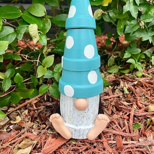 May include: A whimsical garden gnome figurine. The gnome has a stacked pot hat painted turquoise with white polka dots. The body is white with a tan nose and feet. The gnome is sitting on mulch.