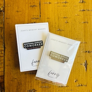 Puede incluir: Dos pines de esmalte sobre tarjetas blancas con el texto "JEAN'S MEMORY MAKERS". Los pines dicen "EMOTIONAL SUPPORT COWORKER PLEASE DO NOT PET" y "funny ENAMEL PIN". Un pin está en una funda de plástico.