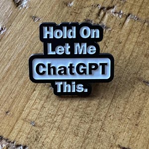 以下が含まれることがあります： 「Hold On Let Me ChatGPT This」のテキストが入った、白黒の長方形のピン。ピンは黒い縁取りと白い文字で構成されています。ピンは木製の表面にあります。