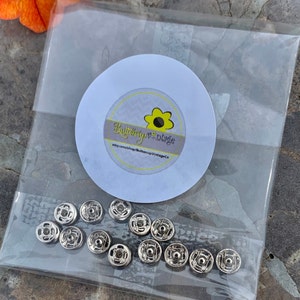 May include: A clear plastic bag containing a set of 12 silver metal snap fasteners. The bag has a white circle sticker with the text "Buttercup Vintage" and a yellow flower graphic.