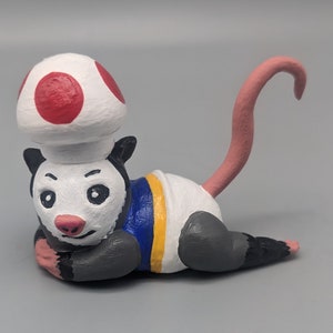 May include: A hand-painted figurine of a possum wearing a red and white mushroom cap and a blue and yellow outfit. The possum has a long, pink tail.