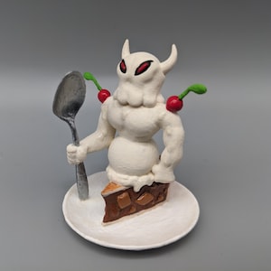 May include: A white, sculpted figure with red eyes and a cherry on its head, holding a silver spoon and standing on a white plate with a chocolate and caramel dessert.