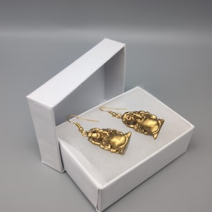 May include: A pair of gold earrings with a small, detailed figure of a woman with a red gemstone in her eye. The earrings are in a white box.