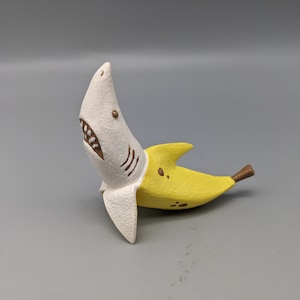 May include: A whimsical sculpture of a banana with a shark head. The shark head is white with brown details and a toothy grin. The banana is yellow with brown spots and a brown stem. The sculpture is set against a gray background.