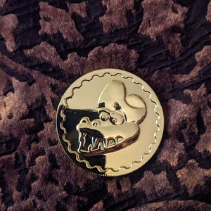 Puede incluir: Una moneda de color dorado con una imagen detallada de la cara de un monstruo gruñendo. La moneda tiene un borde festoneado.