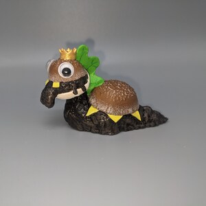 May include: A small, brown and green figurine of a burger with a crown on its head. The burger is on a brown base and has a green leaf on its side.