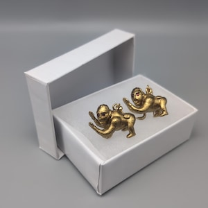 May include: Two gold-colored monkey figurines with red eyes, sitting in a white box. The monkeys are in a crouching position with their arms outstretched.