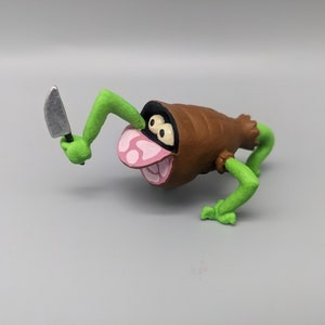 May include: A green and brown figurine of a creature with a knife. The creature has a brown sausage-like body, green arms and legs, and a pink mouth with white teeth.