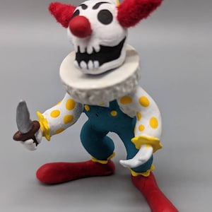 May include: A small, hand-painted clown figurine with a skull face, red hair, a yellow and white hat, a white collar, a blue and yellow polka dot outfit, and a knife in its hand.