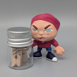 May include: A small glass jar with a silver lid containing two wooden clothespins. A small red-hooded figure with blue jeans and white shoes stands next to the jar.
