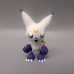 May include: A white, 3D printed figurine of a cartoon character with large, blue eyes, a yellow crescent moon on its forehead, and purple gloves and boots.