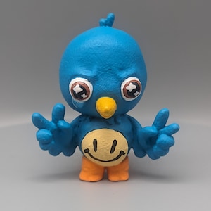 May include: A blue bird figurine with a yellow smiley face on its chest. The bird has large, round eyes with white pupils and is holding its wings out in a peace sign gesture. The bird is painted with a blue body, orange legs, and a yellow beak.