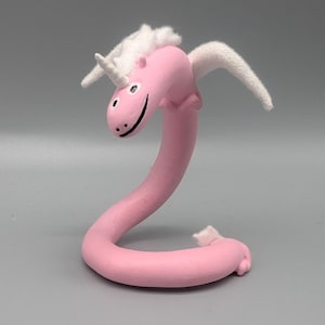 May include: A whimsical pink unicorn sculpture with a curved body, white mane, wings, and horn. The unicorn has a smiling face with black eyes and a black smile. The sculpture is on a gray background.