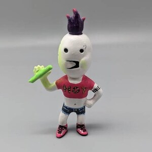May include: A white figurine with a purple mohawk, wearing a red crop top with the word "HOT" on it, blue denim shorts, and pink shoes. The figurine is holding a green object in its right hand.