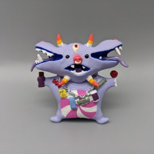 May include: A purple and blue monster figurine with two mouths, horns, and candy-themed accessories. The monster is wearing a pink and white candy-striped shirt and a silver chain with charms.