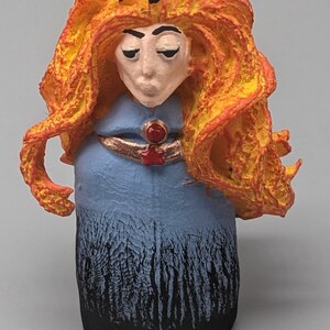 May include: A hand-crafted ceramic figurine of a woman with long, fiery orange hair, wearing a blue dress with a black and white pattern, and a gold and red crown.