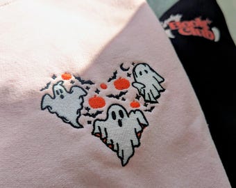 May include: A light pink sweatshirt with a heart-shaped design of embroidered ghosts, bats, and pumpkins. A black sweatshirt with the text "Boo Club" in orange is visible in the background. The sweatshirts are likely for Halloween.