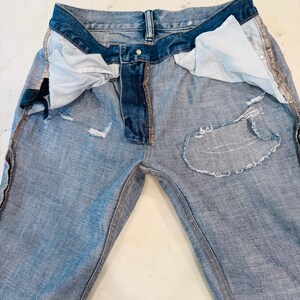 Puede incluir: Vaqueros de mezclilla azul claro con detalles desgastados, incluyendo parches rasgados y bordes deshilachados. Los vaqueros tienen un dise&ntilde;o cl&aacute;sico de cinco bolsillos, cierre de bot&oacute;n y cremallera, y una cinturilla azul oscuro.