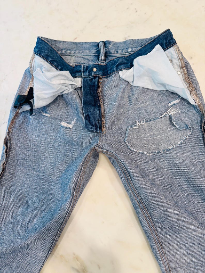 Puede incluir: Vaqueros de mezclilla azul claro con detalles desgastados, incluyendo parches rasgados y bordes deshilachados. Los vaqueros tienen un dise&ntilde;o cl&aacute;sico de cinco bolsillos, cierre de bot&oacute;n y cremallera, y una cinturilla azul oscuro.