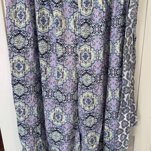 K&ouml;nnte beinhalten: Eine Hose mit Muster in Lila, Gelb und Schwarz, die an einem Kleiderb&uuml;gel h&auml;ngt. Die Hose hat ein sich wiederholendes geometrisches Design und einen kontrastierenden Saum. Der Stoff scheint leicht und f&uuml;r w&auml;rmeres Wetter geeignet zu sein.