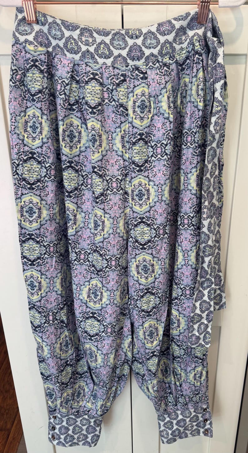 K&ouml;nnte beinhalten: Eine Hose mit Muster in Lila, Gelb und Schwarz, die an einem Kleiderb&uuml;gel h&auml;ngt. Die Hose hat ein sich wiederholendes geometrisches Design und einen kontrastierenden Saum. Der Stoff scheint leicht und f&uuml;r w&auml;rmeres Wetter geeignet zu sein.