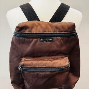 May include: A brown backpack with black zippers and straps. The backpack has a small front pocket and a top zipper. The brand name "kate spade NEW YORK" is visible on a small black tag.