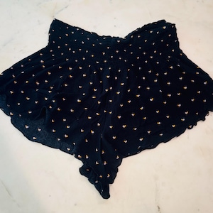 May include: Navy blue shorts with a repeating pattern of small yellow hearts. The shorts have a gathered waistband and a flowing, scalloped hemline. The fabric appears lightweight and suitable for warm weather.