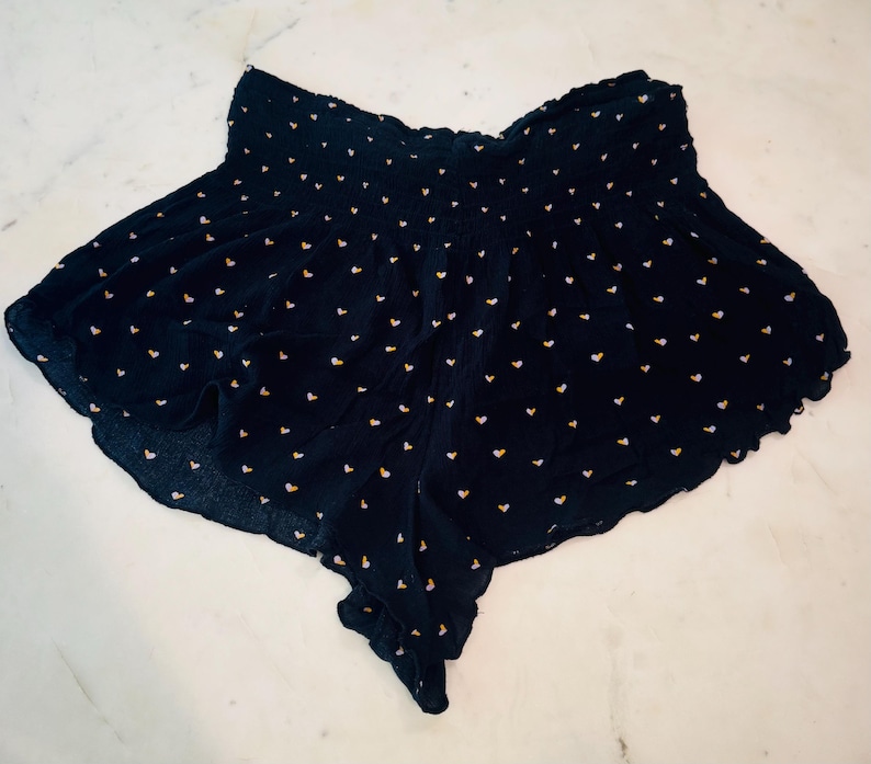 May include: Navy blue shorts with a repeating pattern of small yellow hearts. The shorts have a gathered waistband and a flowing, scalloped hemline. The fabric appears lightweight and suitable for warm weather.