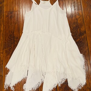 Puede incluir: Vestido blanco de tirantes finos con cuello en V y dobladillo asim&eacute;trico. El vestido presenta detalles de flecos en el dobladillo y las costuras, creando un estilo bohemio fluido.