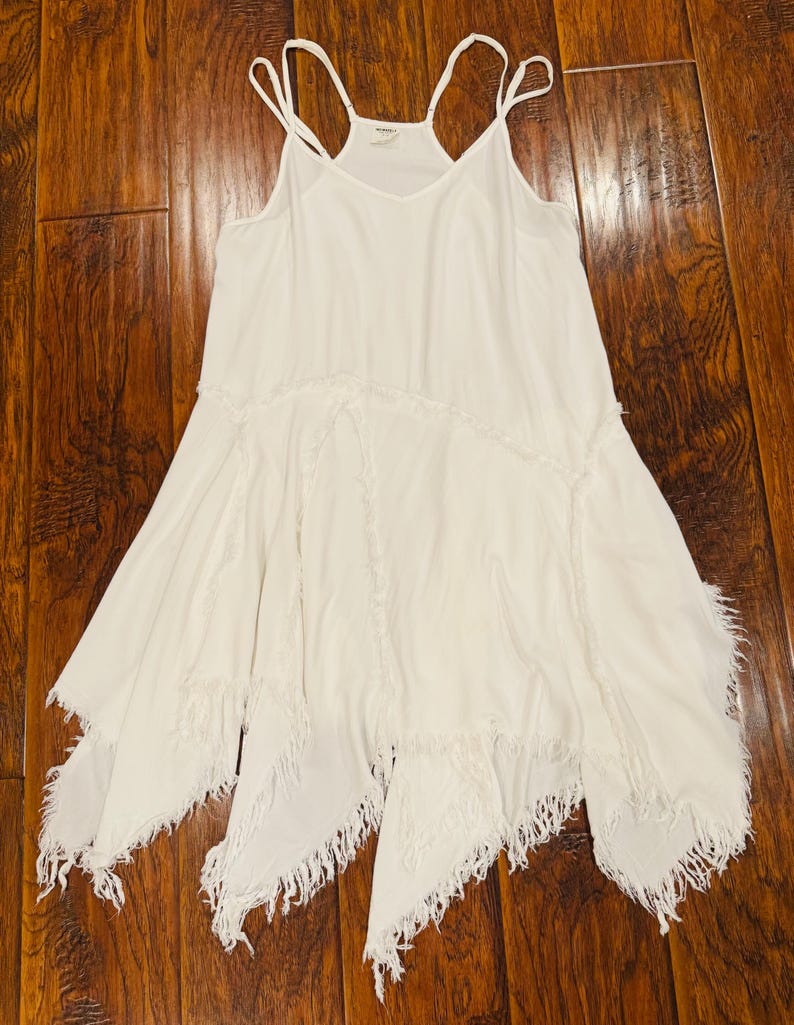 Puede incluir: Vestido blanco de tirantes finos con cuello en V y dobladillo asim&eacute;trico. El vestido presenta detalles de flecos en el dobladillo y las costuras, creando un estilo bohemio fluido.