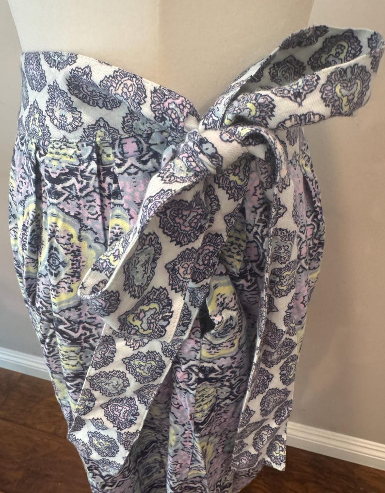 K&ouml;nnte beinhalten: Wickelrock mit Paisley-Muster in Wei&szlig;, Lavendel, Gelb und Schwarz. Der Rock wird mit einem passenden Stoffg&uuml;rtel in der Taille gebunden, wodurch ein geschichteter Effekt entsteht. Der Stoff wirkt weich und flie&szlig;end.