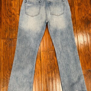 Puede incluir: Vaqueros de mezclilla azul claro con un aspecto desgastado, con &aacute;reas descoloridas y dobladillos deshilachados. Los vaqueros tienen dos bolsillos traseros y un dise&ntilde;o cl&aacute;sico de cinco bolsillos. La tela parece ser una mezclilla de peso medio.