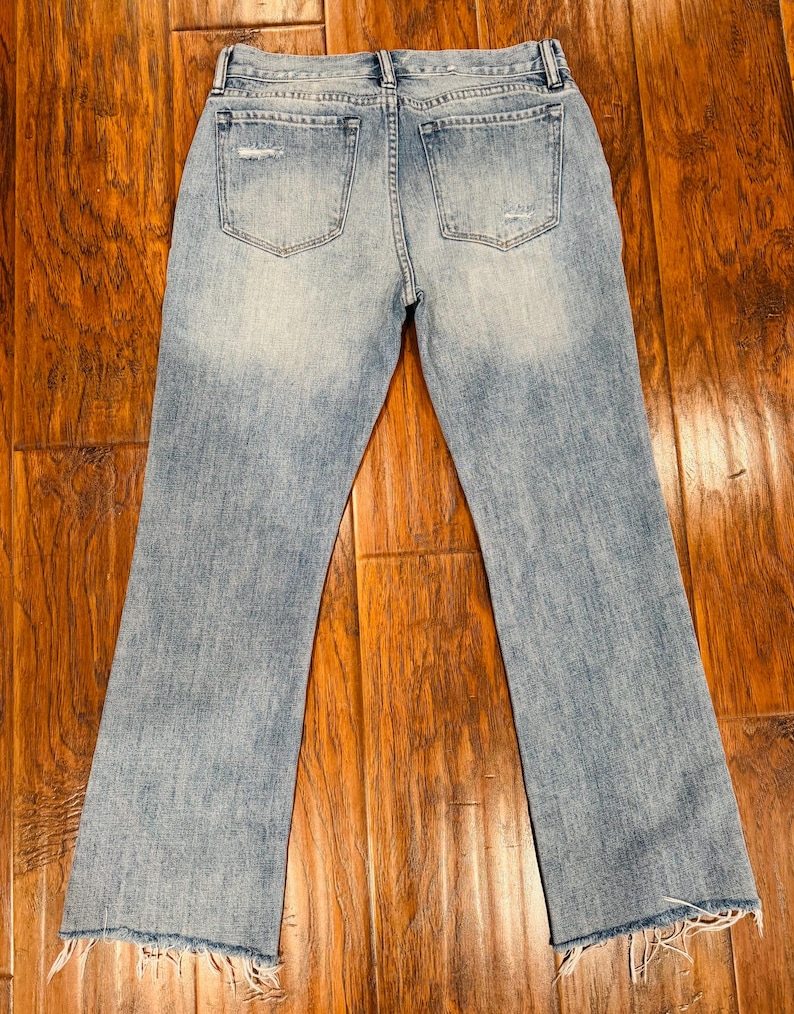 Puede incluir: Vaqueros de mezclilla azul claro con un aspecto desgastado, con &aacute;reas descoloridas y dobladillos deshilachados. Los vaqueros tienen dos bolsillos traseros y un dise&ntilde;o cl&aacute;sico de cinco bolsillos. La tela parece ser una mezclilla de peso medio.