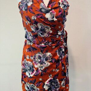 K&ouml;nnte beinhalten: Ein tr&auml;gerloses Kleid mit floralem Muster in einem leuchtenden Orangeton. Das Kleid hat gro&szlig;e, stilisierte Blumen in Wei&szlig;-, Lila- und Rosat&ouml;nen mit dunkelvioletten Bl&auml;ttern. Der Stoff ist gerafft und ger&uuml;scht und schafft eine schmeichelhafte Silhouette.