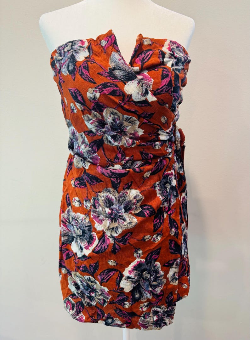 K&ouml;nnte beinhalten: Ein tr&auml;gerloses Kleid mit floralem Muster in einem leuchtenden Orangeton. Das Kleid hat gro&szlig;e, stilisierte Blumen in Wei&szlig;-, Lila- und Rosat&ouml;nen mit dunkelvioletten Bl&auml;ttern. Der Stoff ist gerafft und ger&uuml;scht und schafft eine schmeichelhafte Silhouette.