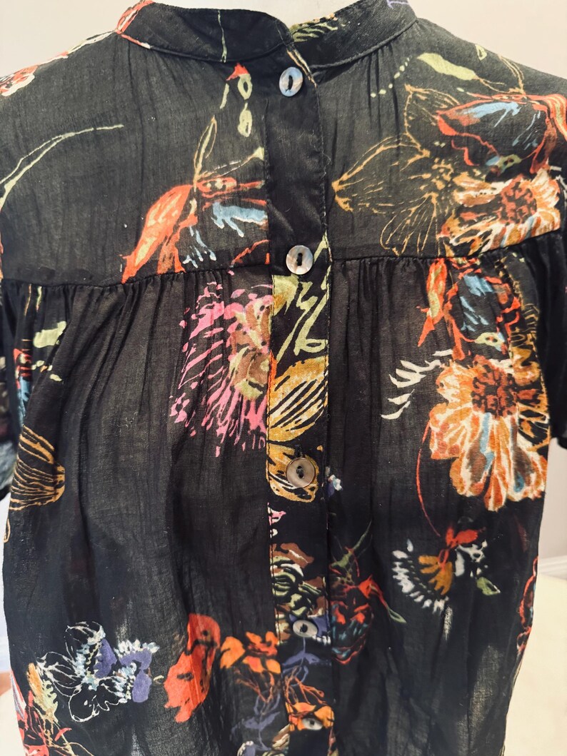 May include: Black button-down shirt with a floral pattern in shades of orange, pink, and blue. The shirt has a mandarin collar and short sleeves. The fabric appears to be lightweight and semi-sheer.