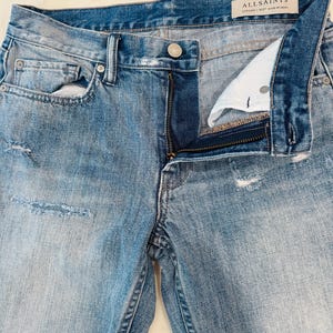 Puede incluir: Vaqueros de mezclilla azul claro con cierre de bot&oacute;n y cremallera. Los vaqueros tienen detalles desgastados y un lavado descolorido. La marca "ALLSAINTS" es visible en una etiqueta.