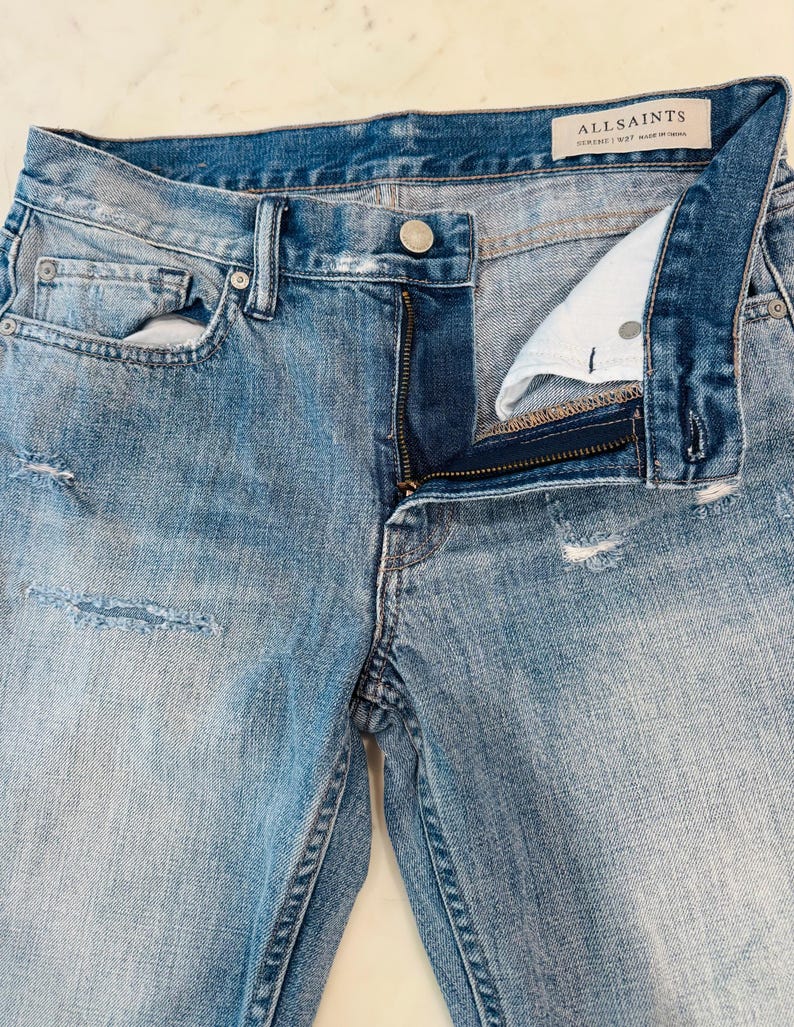 Puede incluir: Vaqueros de mezclilla azul claro con cierre de bot&oacute;n y cremallera. Los vaqueros tienen detalles desgastados y un lavado descolorido. La marca "ALLSAINTS" es visible en una etiqueta.