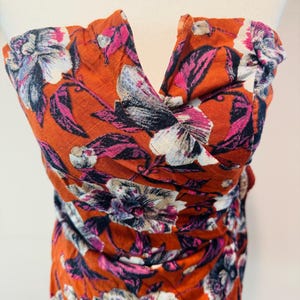 K&ouml;nnte beinhalten: Ein orangefarbenes tr&auml;gerloses Oberteil mit floralem Muster. Die Blumen sind wei&szlig;, rosa und lila. Der Stoff ist gerafft und ger&uuml;scht, wodurch ein strukturierter Look entsteht. Das Oberteil wird auf einer Schaufensterpuppe pr&auml;sentiert.