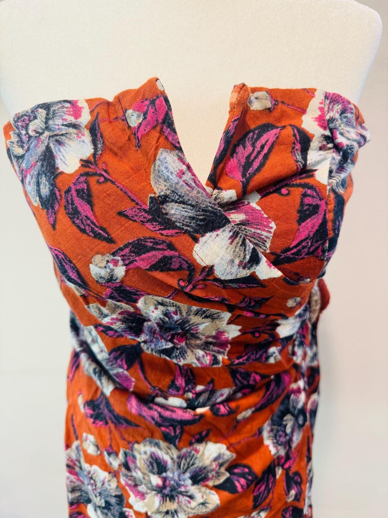 K&ouml;nnte beinhalten: Ein orangefarbenes tr&auml;gerloses Oberteil mit floralem Muster. Die Blumen sind wei&szlig;, rosa und lila. Der Stoff ist gerafft und ger&uuml;scht, wodurch ein strukturierter Look entsteht. Das Oberteil wird auf einer Schaufensterpuppe pr&auml;sentiert.