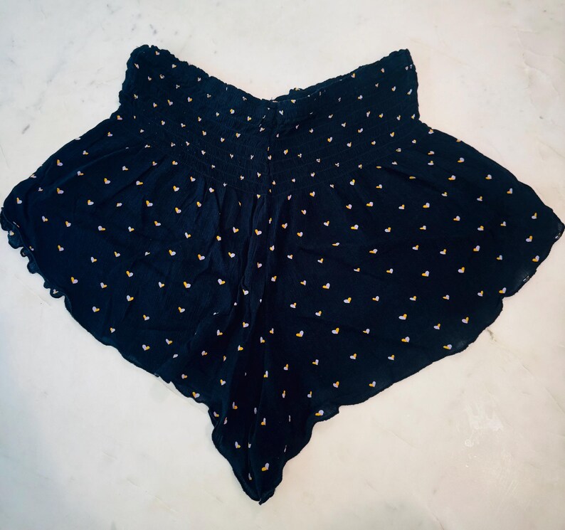 May include: Navy blue shorts with a high, elasticized waist and a scalloped hem. The shorts are covered in tiny white and yellow heart-shaped patterns. The fabric appears to be lightweight and flowy.