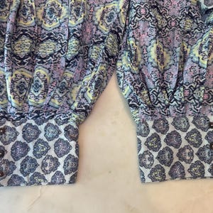 K&ouml;nnte beinhalten: Nahaufnahme eines Kleidungsst&uuml;cks mit farbenfrohem Muster. Der Stoff weist eine Mischung aus Blau-, Lila- und Gelbt&ouml;nen auf. Die Manschetten haben ein kontrastierendes wei&szlig;es und blaues Paisley-Muster und sind mit braunen Kn&ouml;pfen befestigt.