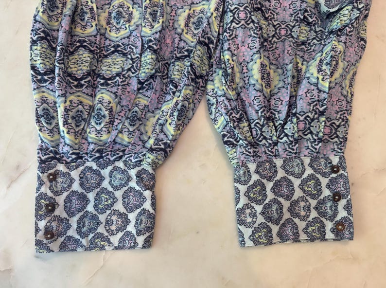K&ouml;nnte beinhalten: Nahaufnahme eines Kleidungsst&uuml;cks mit farbenfrohem Muster. Der Stoff weist eine Mischung aus Blau-, Lila- und Gelbt&ouml;nen auf. Die Manschetten haben ein kontrastierendes wei&szlig;es und blaues Paisley-Muster und sind mit braunen Kn&ouml;pfen befestigt.