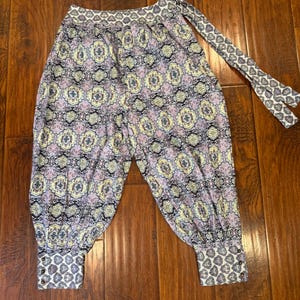 K&ouml;nnte beinhalten: Eine Hose mit Muster im Harem-Stil mit einem sich wiederholenden Blumenmuster in Lila, Gelb und Schwarz. Die Hose hat einen gemusterten Bund und Manschetten sowie eine passende Krawatte.