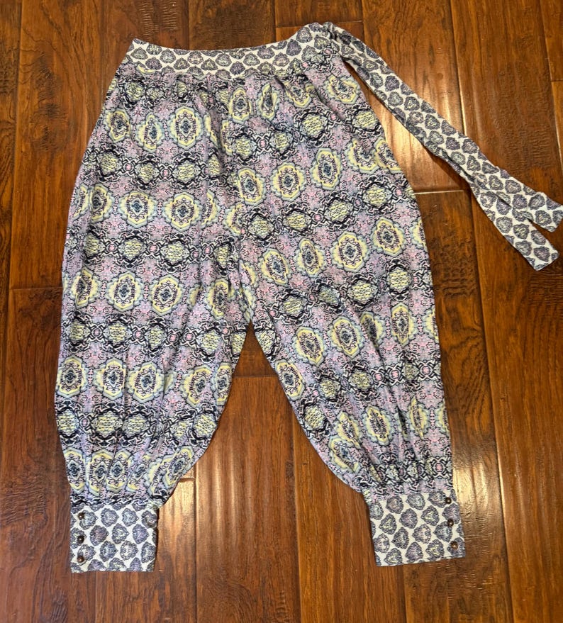 K&ouml;nnte beinhalten: Eine Hose mit Muster im Harem-Stil mit einem sich wiederholenden Blumenmuster in Lila, Gelb und Schwarz. Die Hose hat einen gemusterten Bund und Manschetten sowie eine passende Krawatte.