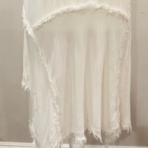Puede incluir: Vestido blanco asim&eacute;trico con bordes deshilachados. El vestido se exhibe sobre un pedestal blanco. La tela parece ligera y fluida, con un dise&ntilde;o en capas y un ribete texturizado y con flecos.